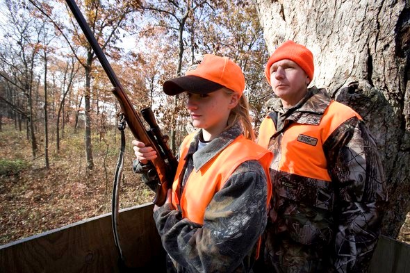 A girl holds her rifle in the hunter position as her father looks on.