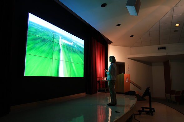 A woman stands on a stage in front of a video wall and sound system