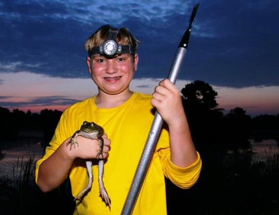A boy in a yellow shirt and a bright headlamp holds a bullfrog and a gig