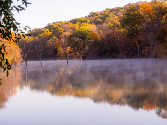 Fall foliage is mirrored in on a glassy lake. Morning fog steams off the water. 
