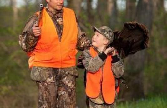 Father and son wearing hunter orange return from a successful turkey hunt. The boy has a turkey over his shoulder.