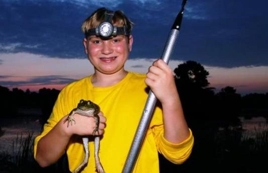 A boy in a yellow shirt and a bright headlamp holds a bullfrog and a gig